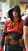 Photo d'un homme portant un costume rouge, noir et doré avec sa capuche sur la tête.