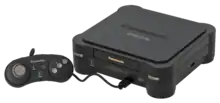 Photo d'une console noire, sur laquelle on trouve l’inscription Panasonic. Une manette est branchée à cette console.