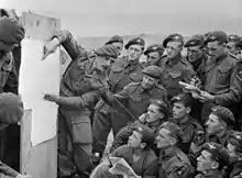 Photo en noir et blanc d'un homme en uniforme parlant à d'autres hommes, assis ou debout tout autour de lui, qui écoutent attentivement le premier en regardant la carte qu'il montre.