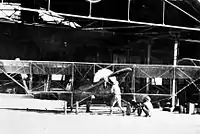 Photographie en noir et blanc d'un biplan monomoteur et de deux hommes devant un grand hangar ouvert