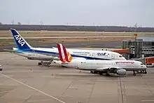 Photo de deux avions à la porte d'embarquement d'un aéroport. Au premier plan, un avion plus petit, aux couleurs blanches, oranges et rouges, cache la moitié d'un autre avion plus grand aux couleurs blanches et bleues, présent à ses côtés.