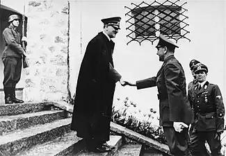 Photo noir et blanc, prise le 9 juin 1941 à Berchtesgaden, en Allemagne. Au centre de la photo, Adolf Hitler, portant un long manteau noir et une casquette militaire, serre la main d’Ante Pavelić (à droite), dirigeant de l’État indépendant de Croatie. Les deux hommes sont de profil, debout, sur les marches de l’escalier menant à l’entrée de la résidence secondaire de Hitler. Un soldat en arme se tient debout, en haut de l’escalier (en haut à gauche de la photo). Au second plan, deux officiers allemands (en bas à droite de la photo) assistent à la scène. L’arrière-plan de la photo est constitué d’un mur blanc percé d’une fenêtre grillagée (au-dessus de la tête du dirigeant croate).