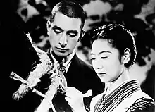Un homme et une femme se tiennent devant le tronc d’un arbre entouré d’une corde, la femme noue un bout de tissu sur la corde (image en noir et blanc).