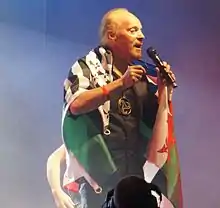 Alan Stivell tenant son micro avec le drapeau sur les épaules.