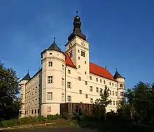 Photo en couleurs (18 août 2005) du château de Hartheim, en Bavière, centre d’euthanasie ouvert pendant la période nazie. La couleur beige très clair des deux façades visibles du château, à l’architecture de type Renaissance, contraste contre le ciel bleu, en arrière-plan, et la verdure au premier plan.