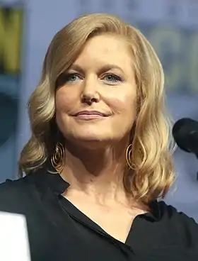 Photo en buste d'une femme souriante en face d'un micro, avec des cheveux longs blonds et ondulés, portant des boucles d'oreilles dorées et habillée d'un chemisier noir.