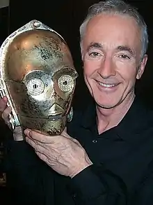 Homme blanc aux cheveux gris et aux yeux bleus tenant la tête d'un robot doré.
