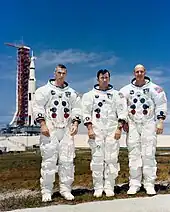 Trois hommes debouts en combinaison spatiale. Fusée sur site de lancement à l'arrière plan.