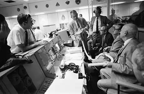 Au centre de contrôle de la mission à Houston, Deke Slayton montre aux responsables du centre et de la mission le bricolage (surnommé « mailbox ») proposé pour éliminer le dioxyde de carbone de l'atmosphère du vaisseau.
