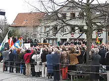 Photographie d'une place publique, avec une foule regardant une estrade