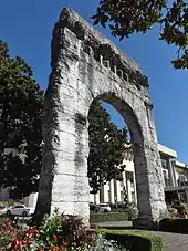 Photographie en couleurs des vestiges d'un arc de triomphe romain ; gra, d bâtiment à l'arrière-plan.