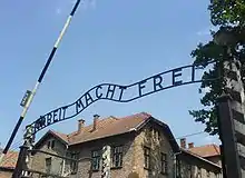 Portail d'entrée d'un camp, avec une barrière levée et une inscription en allemand en fer forgé