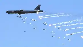 Passage à basse altitude d'un B-52 de couleur très sombre. Sa soute à bombes est ouverte et une nuée de bombes est en train de sortir par cette ouverture.