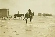2 chevaux montés, dont 1 tire  une cabine cubique à 4 roues vers une rangée de cabines semblables déjà placées au large de la plage.