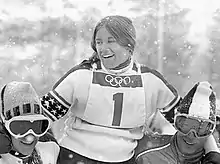 Photographie en noir et blanc montrant une skieuse avec le dossard no 1 portée en triomphe par deux autres personnes.
