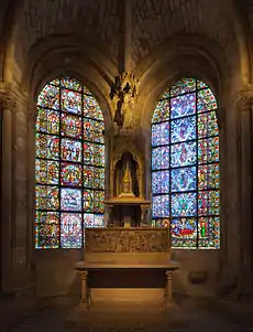 Photographie contemporaine d'un reliquaire de pierre situé entre les vitraux colorés d'une petite chapelle.