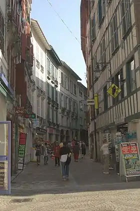 Photographie d’une rue étroite, s'incurvant vers la droite et bordée d’immeubles de 3 étages, aux façades blanches à pans de bois peints.