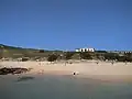 Plage au premier plan, avec deux petites embarcations, quelques touristes puis à l'arrière plan, une maison et un autre bâtiment sur la droite ; quelques arbres.
