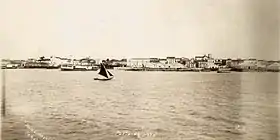 Photographie d'un plan d'eau avec un petit voilier au premier plan et des bâtiments le long du rivage en arrière-plan