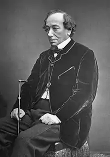 Portrait d'un homme âgé à l'air fatigué assis sur une chaise. Il arbore un petit bouc et a les cheveux bouclés rabattus sur le côté. Il porte une veste noire en velours, un monocle en pendentif et tient une canne.