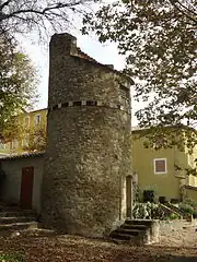 Photo prise en sous-bois. Le pigeonnier est une tour construite en moellons apparents et à toit incliné, avec un rang de carreaux émaillés de couleurs qui en fait le tour à 4 m de haut.