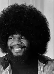 Photographie en noir et blanc d'un homme aux cheveux frisés, portant la barbe, souriant et regardant vers sa droite.