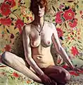 Portrait de face d'une femme nue assise sur un canapé fleuri, visage à moitié dans l'ombre, jambes repliées et bras droit cachant en partie le sexe