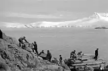 Photo noir et blanc de dix personnes transportant du matériel sur un pente rocheuse dans un paysage de glace.