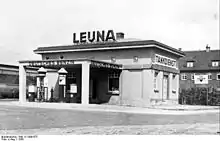 Photographie en noir et blanc montrant une station-service. Le mot « LEUNA » apparaît au-dessus de l'immeuble, alors que les expressions « Deutches Benzin » et « Tankdienst » décorent certaines parties de l'immeuble.