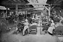 Photographie en noir et blanc d’une immense salle que les verrières du plafond, qui repose sur des poutres et des pilastres, éclairent chichement. À perte de vue se répètent les rangées de mêmes machines actionnées par des femmes assises, en coiffes et longs tabliers blanc. De rares hommes déambulent dans les allées, surveillant…