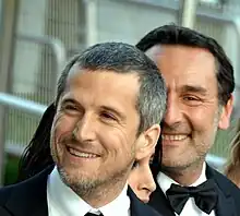 Deux hommes en smoking, souriants, l'un à moitié caché derrière l'autre.