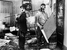 Photographie en noir et blanc d'un homme portant un balai sur son épaule et frappant par mégarde un homme placé derrière lui.