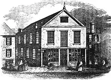 Une gravure en noir et blanc représentant une scène de rue avec, au centre, une église de 2,5 étages avec des fenêtres à meneaux.