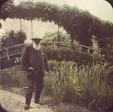 Photographie coloriée d'un vieillard à barbe blanche devant un petit pont courbe entouré de verdure.
