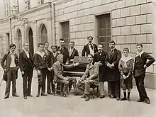 Vue en noir et blanc de 11 hommes et une femme debout derrière un piano devant lequel sont assis 2 hommes.