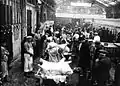 Concours du meilleur ouvrier boucher aux abattoirs de Vaugirard en 1932 : sous une halle des participants découpent sur des sortes de tréteaux des carcasses de moutons entourés par un public, en costume de ville ou de travail. Le sol est couvert de sang et de débris de laine.