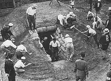 Photographie en noir et blanc montrant des hommes et des femmes construisant un monticule en terre avec une ouverture sur le côté. L'entrée est bordée de sacs de sable.