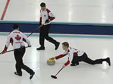 Photographie de trois curleurs, l'un à droite en position allongée, genou plié, tenant dans la main droite une pierre, dans la main gauche un balai, les deux autres debout portant un balai.