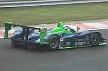Photographie d'une voiture de sport-prototype bleue, verte et blanche, sur une piste, vue de trois-quarts arrière.