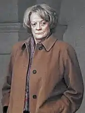 Photo en demi-profil (gauche) d'une femme au visage ridé, qui se tient adossée à une colonne en pierre avec les mains dans les poches. Ses cheveux sont grisonnants, coupés «&nbsp;au bol&nbsp;». Elle porte un trench-coat marron et son regard est concentré sur quelque chose situé à la droite du photographe.