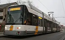 La photo couleur montre une rame grise en blanche rayée de jaune-orangé dans une rue pavée.