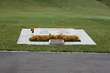 Plaque de marbre portant les noms de John et Olive Diefenbaker se trouvant au milieu d'une pelouse et entourée de fleurs jaunes et d'une petite plaque de bronze.