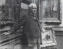 Paul Durand-Ruel debout s'appuie contre un mur de sa galerie où plusieurs tableaux sont accrochés.