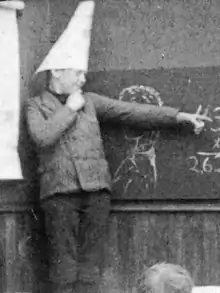 Photographie d'un élève portant un bonnet d'âne devant un tableau noir