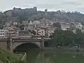 Pont en pierre de taille, dont deux des trois arches sont visibles, l'arche centrale couvre la rivière verdâtre (la Koura), celle de gauche une des berges sur laquelle circulent des automobiles, à l'arrière-plan un bâti dense inclut des immeubles de teintes variées : blanc, rose, gris devant des murailles de teinte grisâtre.