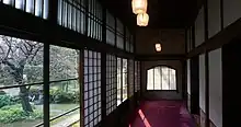 Un couloir de bois en enfilade, portes coulissantes en bois et papier sur la droite, fenêtres sur la gauche. tapis rouge au sol. À l’extérieur par les fenêtres, vue sur un jardin avec un cerisier.