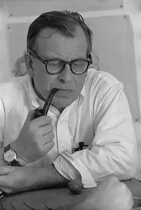 Portrait noir et blanc d’un homme en chemise blanche accoudé à une table et fumant la pipe.