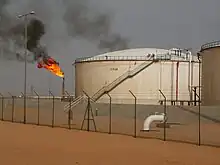 Au premier plan, un grillage est installé sur le sable, devant deux bâtiments circulaires contenant des hydrocarbures, à droite. En arrière-plan à gauche se dresse une torchère enflammée.
