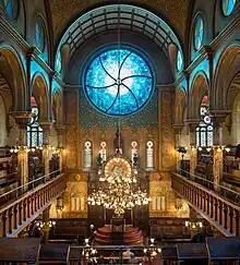 Une grande salle vue de haut. Une grande coupole avec au centre un vitrail bleu. Sur les cotés, des balcon. Vers le bas, un chandelier derrière lequel apparait le sol.