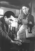 Au premier plan un homme (Ken Uehara), une écharpe autour du cou et l'air sombre, est accroupi et se réchauffe les mains à un brasero, en arrière plan, une femme (Kinuyo Tanaka) pauvrement vétue d'un tablier et d'un gilet, porte un sac en osier et une grande bouteille en verre (image en noir et blanc).